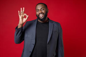 Wall Mural - Smiling cheerful funny young african american business man 20s wearing classic jacket suit standing showing OK gesture looking camera isolated on bright red color wall background studio portrait.