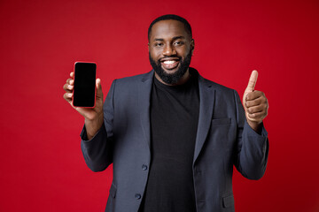 Smiling young african american business man 20s in classic jacket suit standing hold mobile cell phone with blank empty screen showing thumb up isolated on bright red color background studio portrait.