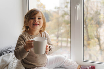 Wall Mural - Child drink cocoa or milk near the window from big white mug. mock up of cup. christmas time