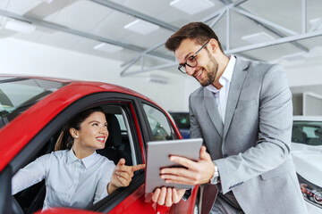 Smiling woman sitting in car and pointing at tablet car seller holding. She is picked right car for her she saw online. Car salon interior.