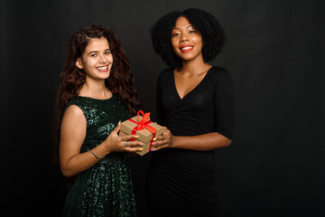 Wall Mural - Two young women of different nationalities are smiling, holding a present for the new year and christmas on a black background. Gift exchange, giving and receiving concept