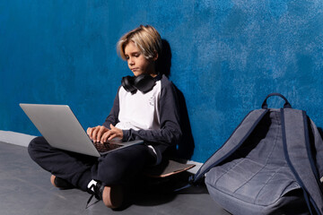 Clever teen boy typing on laptop studying preparing for test exam doing homework, school exercises, sitting on skateboard with backpack. Schoolboy learning assignment. Teen education concept.