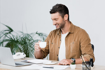 Wall Mural - Cheerful businessman with coffee cup looking at laptop while sitting at workplace with blurred plant on background