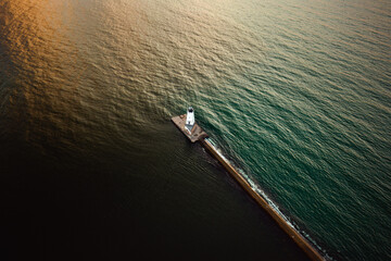Wall Mural - Incredible look down aerial photograph of the Ludington Breakwater Lighthouse sitting at the end of a concrete pier separating dark water from green or blue water lit up by the sun as it begins to set