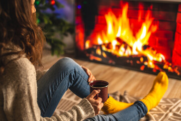 Wall Mural - Girl in a sweater and socks is sitting next to the fireplace with cup of beverage. Concept of creating a cozy atmosphere