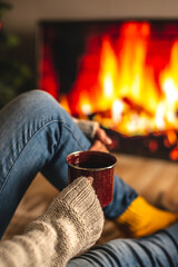 Wall Mural - Girl in a sweater and socks is sitting next to the fireplace with cup of beverage. Concept of creating a cozy atmosphere