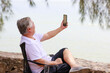 © sutthichai - Asian senior male using mobile phone for video call communication with his family while vacation next to the beach.