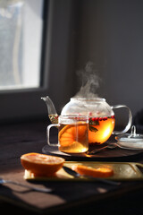 Hot tea with orange and berries in a glass teapot and in a glass mug next to the window in the morning. Sliced orange on a board on a dark background.