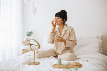 Wall Mural - smiling woman applying face cream sitting on bed