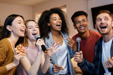 Sing it. Enthusiastic group of friends singing with microphone while playing karaoke at home, standing in the modern apartment