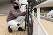 © Andrii Lysenko - Caucasian male Thief in balaclava and hood breaks the lock on a bicycle in the street during the day. Closed face and hacking. Stealing bicycles.