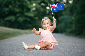 Cute adorable Caucasian baby girl waving Australian flag. Smiling child sitting on street road in park celebrating Australia Day holiday. Celebration of national Australia Day in January outdoors.