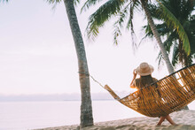 A Young Woman In Straw Hat Sitting In A Hammock Swinging Between A Palm Trees On The Overseas Island Sand Beach At Sunrise Time. Koh Samui, Thailand. Exotic Vacation Concept Image.