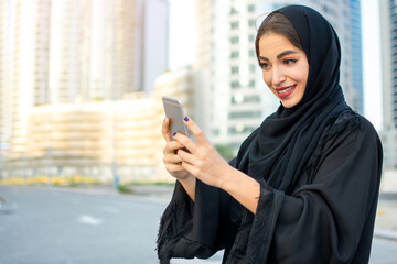 Wall Mural - Portrait of beautiful smiling young Arabic woman in traditional abaya clothes using smartphone on the city street.