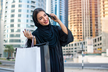 Arab woman in Abaya talking on mobile phone while holding shopping bags and walking on city street.