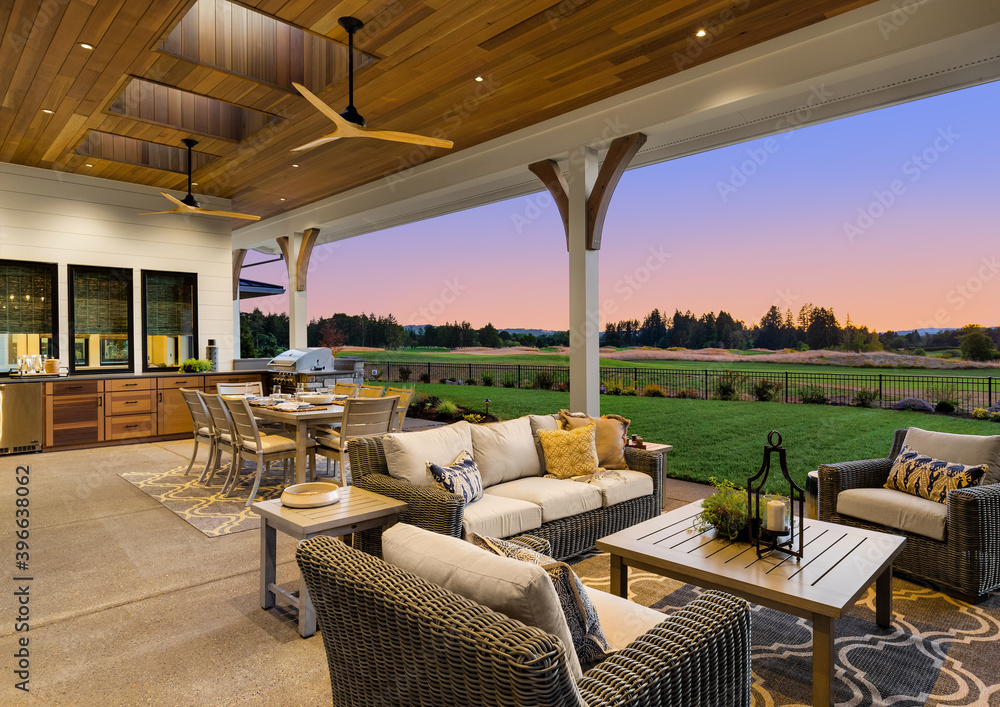 Luxury home exterior at sunset: Outdoor covered patio with kitchen, barbecue, dining table, and seating area, overlooking grass field and trees. - obrazy, fototapety, plakaty 