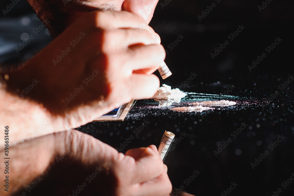 Close up of young drug dealer sniffing drug through rolled one hundred dollar bill from phone screen. Male addict passed out after cocaine overdose on table. Addiction and substance abuse concept