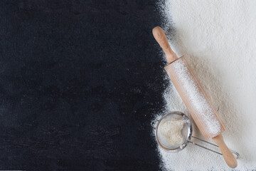 Sifted wheat flour over black surface with rolling pin and sieve top view.