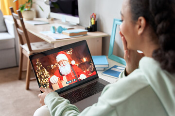 Happy african american teen girl talking to virtual santa claus on video conference online call chat sending Chrtistmas present celebrating remote xmas using laptop computer sitting on bed at home.