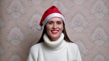 Young Cute Smiling Woman In Christmas Red Cap And White Sweater, Send Air Kiss Into The Camera On Background Of Vintage Wall Wallpaper. Christmas Concept.