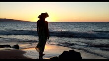 Silhouette Of Stylish Woman Wearing Dress And Hat Standing In Front Of Setting Sun At Sandy Hawaii Beach In Tight Shot. Gentle Waves Rolling To Wet Sand Shoreline With Small, Jagged Lava Rocks.