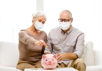 Wall Mural - savings, health and old age concept - senior couple wearing face protective medical mask for protection from virus disease putting coin into big piggy bank