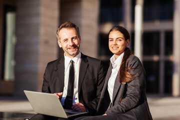 Wall Mural - Portrait of two business peope, middle aged man and young woman in suits, smiling at camera while sitting on bench outdoors and discussing project on laptop