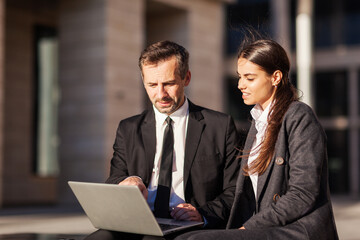 Wall Mural - Middle aged mentor explaining presentation on laptop to young businesswoman sitting on bench outdoors