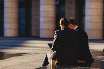 Wall Mural - Back view of business couple in suits sitting on bench outdoors and working on laptop computer together