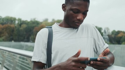 Poster - A smiling african american man is using his smartphone standing on the city bridge