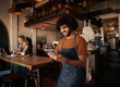 Male waiter with curly hair working in cafe wearing apron with using digital tablet with female working in background on laptop