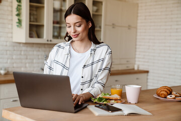 Beautiful smiling woman working with laptop while having breakfast