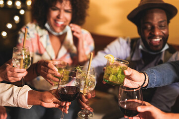 Happy multiracial friends cheering with cocktails while wearing surgical face masks under chin for coronavirus - Home party outdoor on patio