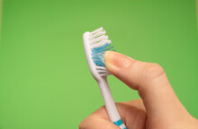 Blue Toothbrush In Woman's Right Hand Against A Green Background With Fingers Guided Through The Bristles