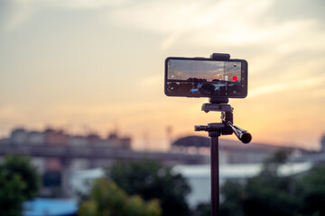 mobile phone mounted on a tripod at full extention replacing a DSLR for a professional shoot to capture a sunset with a metro station and train arriving as seen on the screen with rest out of focus