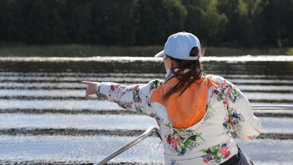 Wall Mural - Tourist caucasian young woman in white cap he points to the shore forest from boat bow deck with railing back view, outdoor active recreation on water in Russia