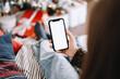 © nikkimeel - Woman holding a smartphone with a white screen mock up, sitting on the bed at home near Christmas tree.