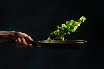 Wall Mural - Professional chef hand throws up frying mix of green vegetables above the pan on dark blue background. Backstage of cooking meal. Frozen motion. Food banner concept.