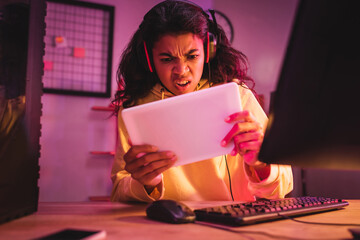 Wall Mural - Angry african american gamer in headset using digital tablet near computer on blurred foreground