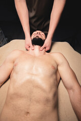 Wall Mural - Male hands of a masseur doing neck massage to a bearded male athlete in a dark room of the massage room