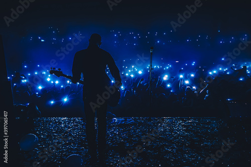 Guitar player in front of crowd on scene in night club. Bright stage lighting, crowded dance floor. Phone lights at concert. Band blue silhouette crowd. People with cell phone lights.