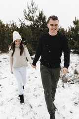 Wall Mural - Young man and woman going on the snow in the pine forest holding each other hands. Love, relationship, winter holidays. Winter couple photo ideas