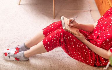 Crop woman sitting on floor at home in solitude writing her dreams or letter for santa claus with her wishes. Female in red pyjamas