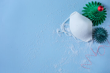 A medical mask and two decorative green Christmas trees and white snow are on a blue background. Free space for text.