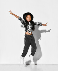 Cute Korean little girl with black hair has fun and jumps with arms outstretched on a white background. Child is wearing a black hat, checkered shirt and jeans. Style for modern kid.