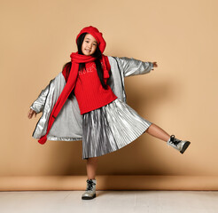 Asian little model girl wearing red beret, scarf, sweater and silver skirt and jacket having fun posing on beige background in studio. Concept of style for modern kid.