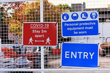 Entrance to a construction site with warning signs attached to a fence. Covid 19 stay 2 metres apart. Personal protective equipment must be worn. Entry.