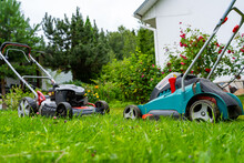 Gasoline And Battery Electric Lawn Mowers In The Garden Against The Backdrop Of A Blooming Garden, Old And New Grass Mowing Technologies