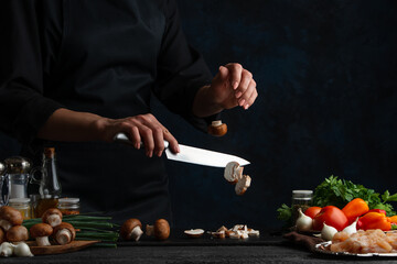 Wall Mural - Professional chef in black uniform cuts with knife mushrooms on the chopped board. Concept of cooking process. Backstage of preparing meat filling for pie on dark blue background. Frozen motion.