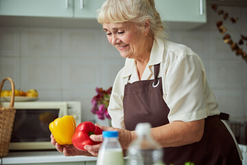 Wall Mural - Cheerful old woman in apron holding bell peppers